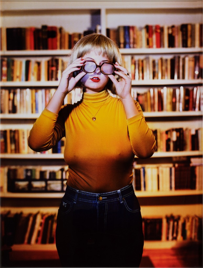 A person in a yellow turtleneck holding glasses in front of bookshelves.